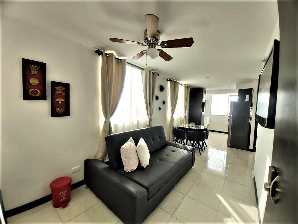 A cozy living area features a dark sofa bed, positioned under a ceiling fan. Natural light floods in through windows adorned with light curtains. A round dining table with chairs is visible, along with a modern kitchen area in the background.