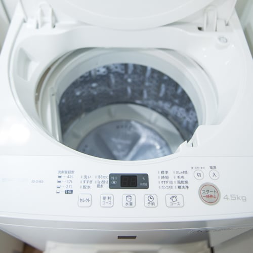 A top-loading washing machine is shown with a transparent lid revealing the drum. The control panel features various settings, clearly labeled. The overall design is modern and compact, suitable for efficient use in limited spaces.