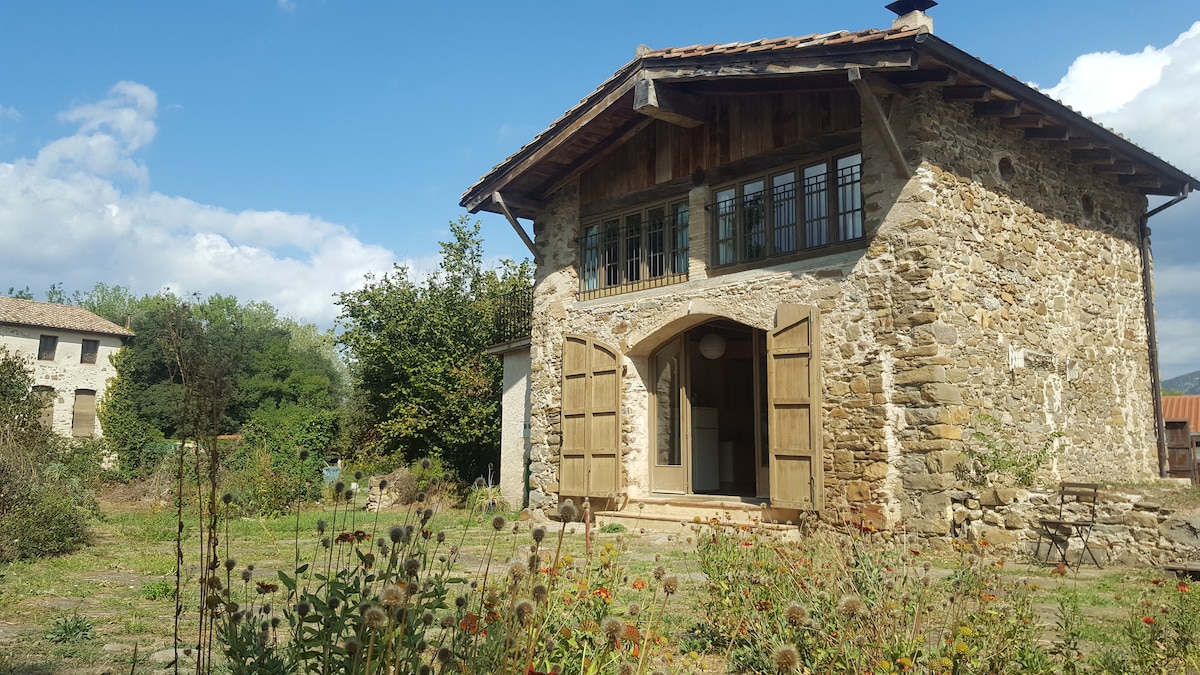 The rustic farmhouse, constructed from local stone, features large wooden doors that open to the inviting entrance. Surrounding gardens are filled with wildflowers, and lush greenery is seen in the background, complementing the peaceful rural setting of La Garrotxa.