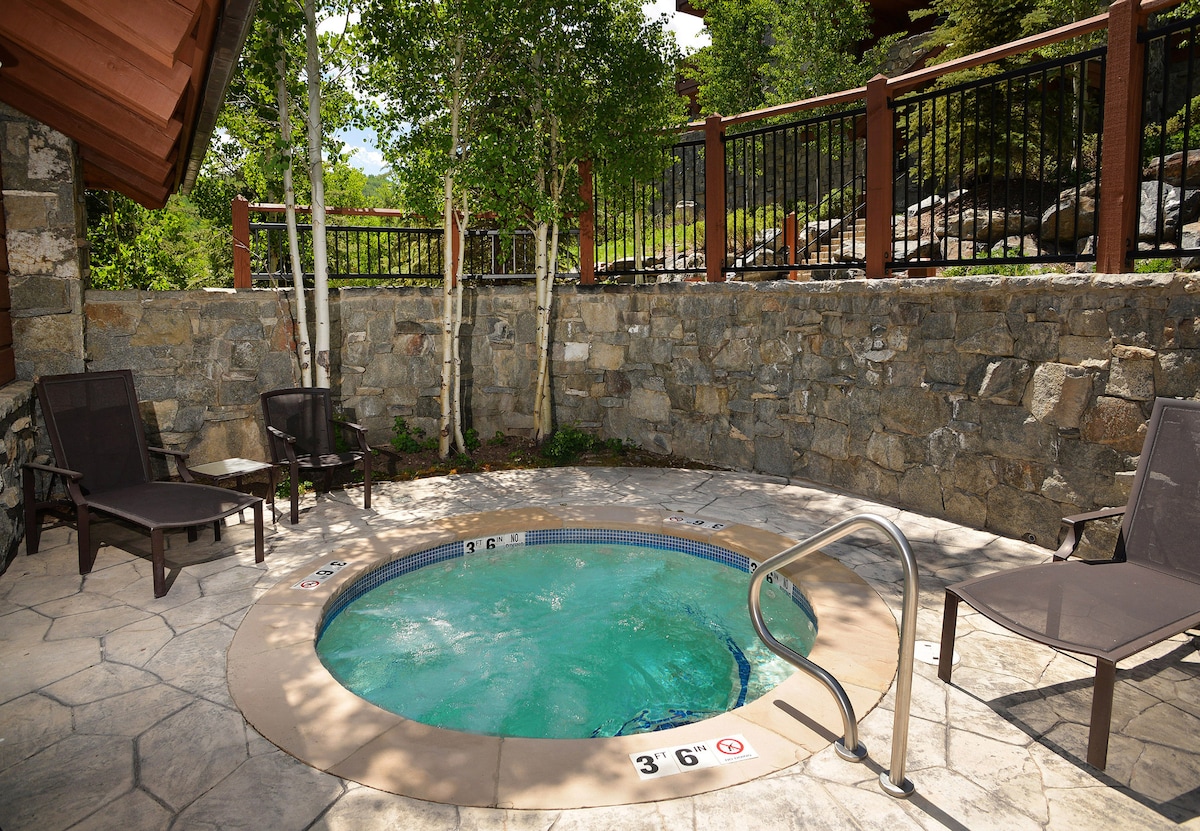 A communal hot tub is set within a stone patio, surrounded by a natural stone wall and lush greenery. Four lounge chairs are positioned nearby, offering a place to relax. Signs indicating water depth are visible, emphasizing safety.