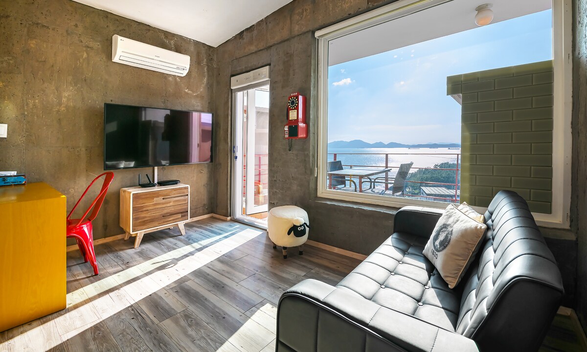 A modern living area showcases a black leather sofa facing a wall-mounted television. A sliding glass door provides access to a patio with scenic views of the water. Natural light fills the space through large windows, complemented by a minimalist wooden table and bright red chairs.