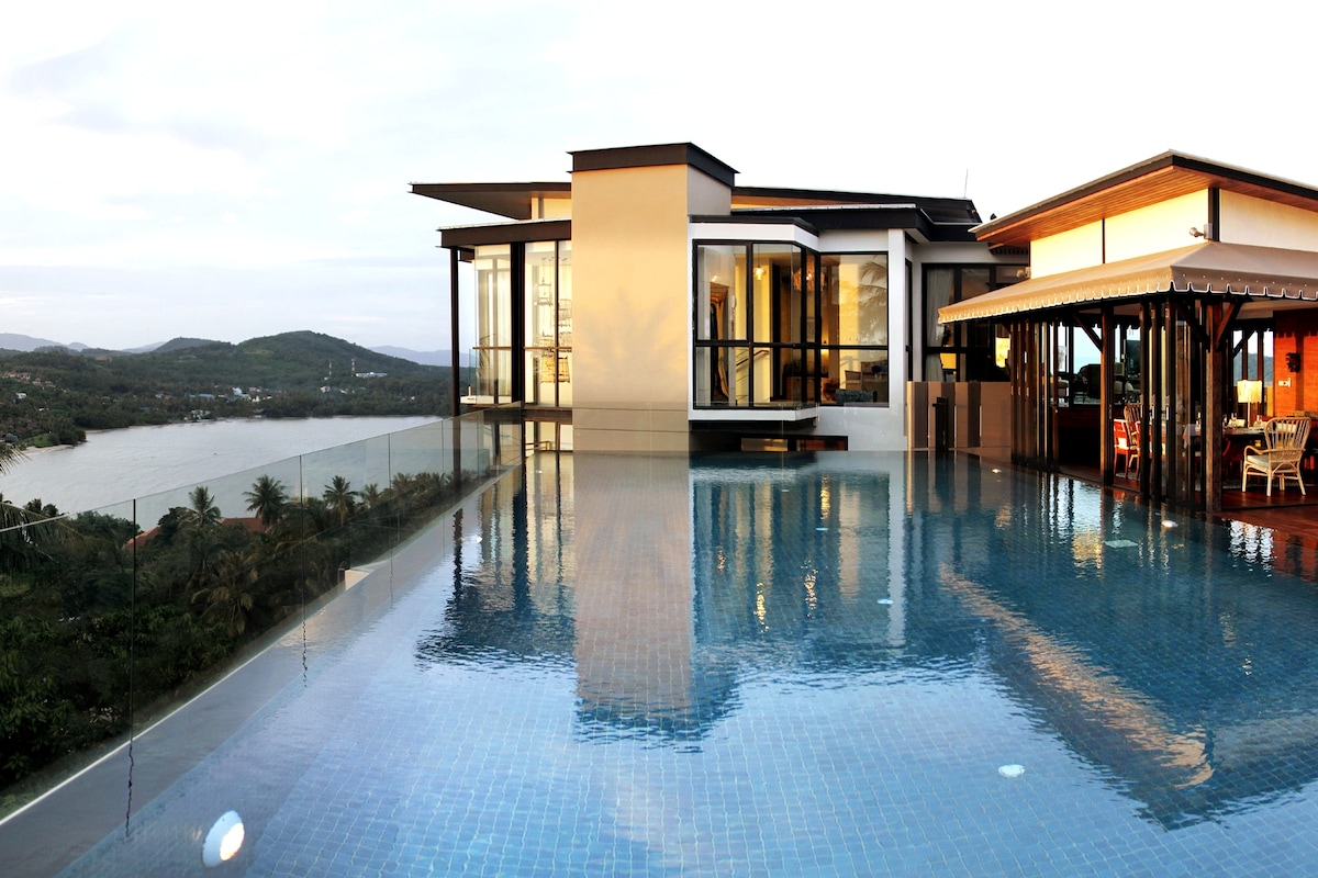 A rooftop infinity pool reflects the sky, seamlessly merging with the tranquil landscape. Modern architecture surrounds the pool, featuring expansive glass walls that offer panoramic views of the Andaman Sea and lush greenery in the distance.