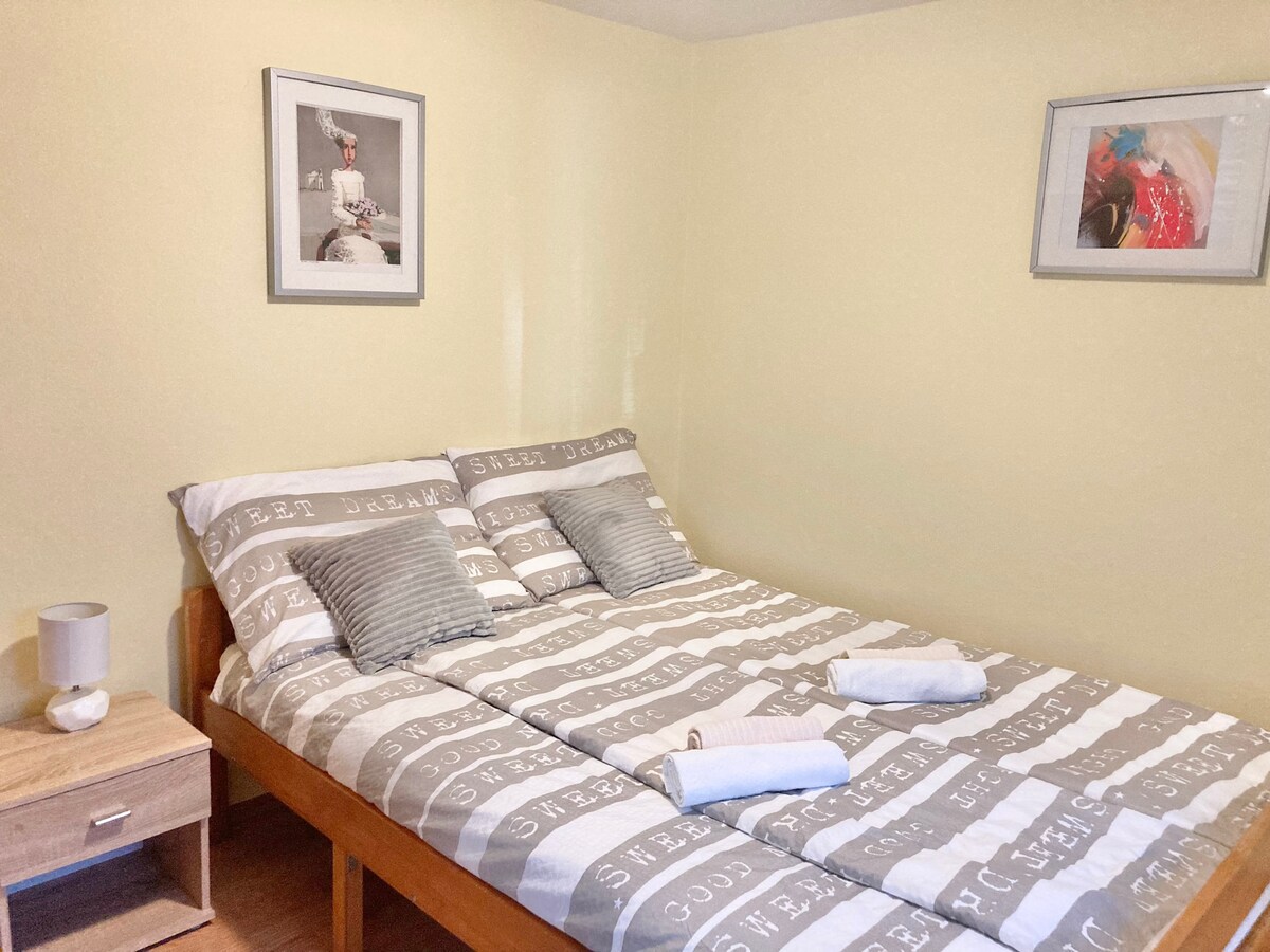 A spacious sleeping area features a wooden bed with neatly arranged bedding in soft grey tones. Two decorative pillows enhance the bed's comfort. Light-colored walls are complemented by framed photographs, and a bedside table holds a small lamp, contributing to a calming ambiance.