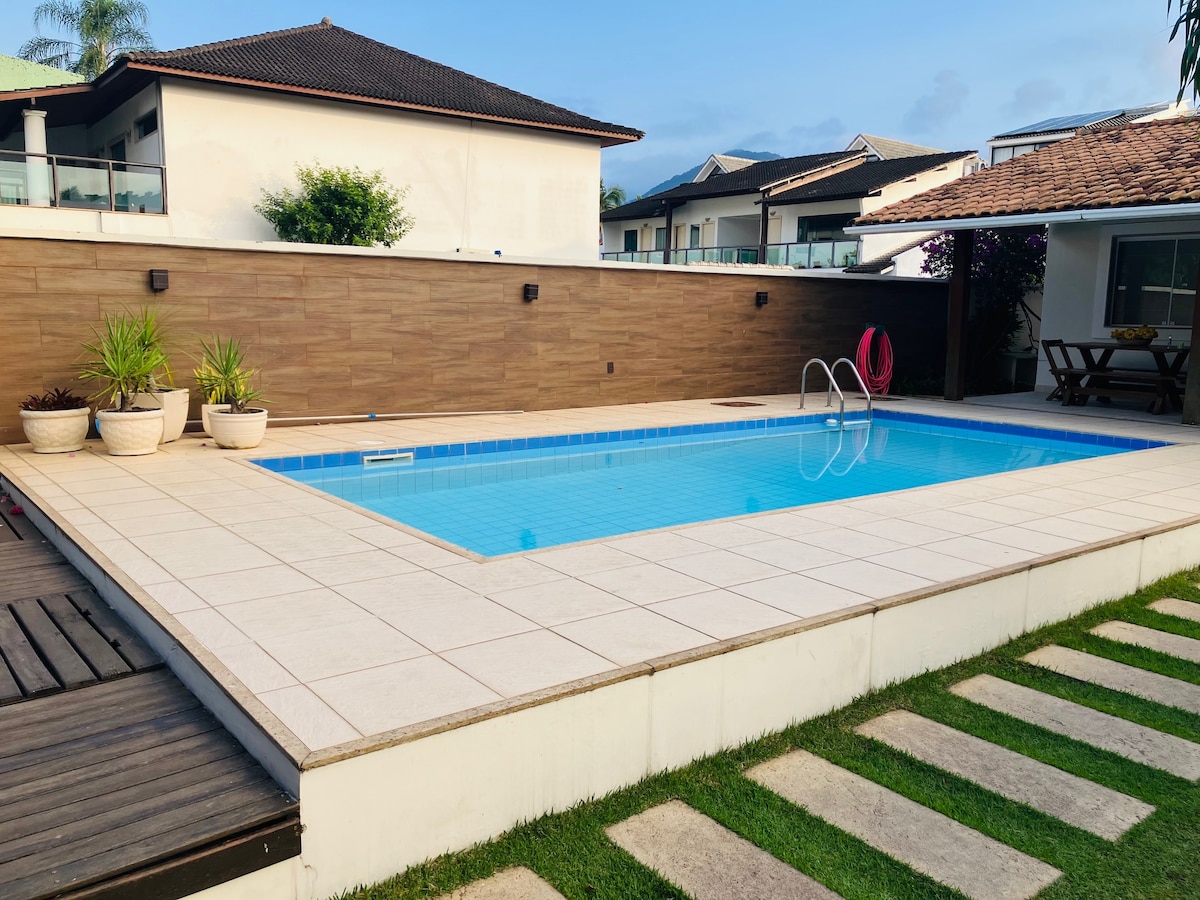 A private swimming pool is framed by a spacious tiled deck, featuring lounge chairs and potted plants. The area is set against a backdrop of the property and neighboring houses, with a grassy border surrounding the pool, creating a serene outdoor space.
