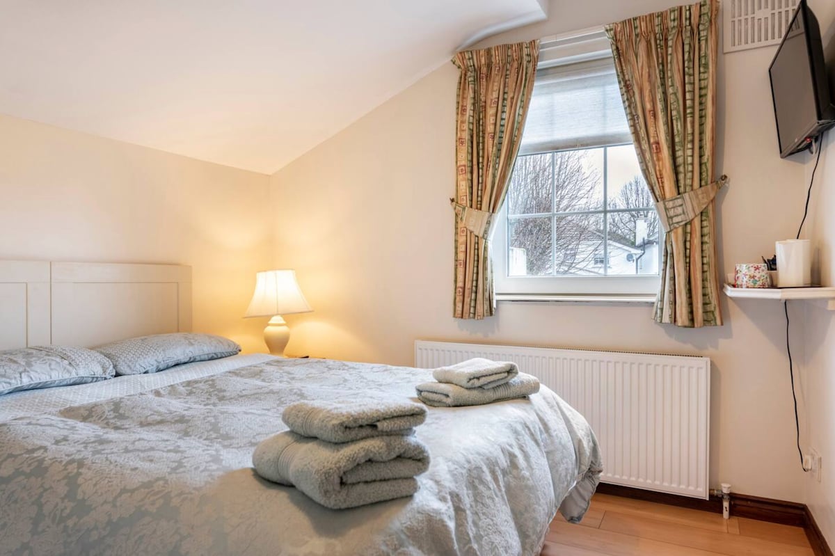 A bright bedroom features a spacious double bed dressed in soft bedding. Natural light flows through a large window, framed by patterned curtains. A bedside lamp stands on a table beside the bed, while towels are neatly arranged at the foot.