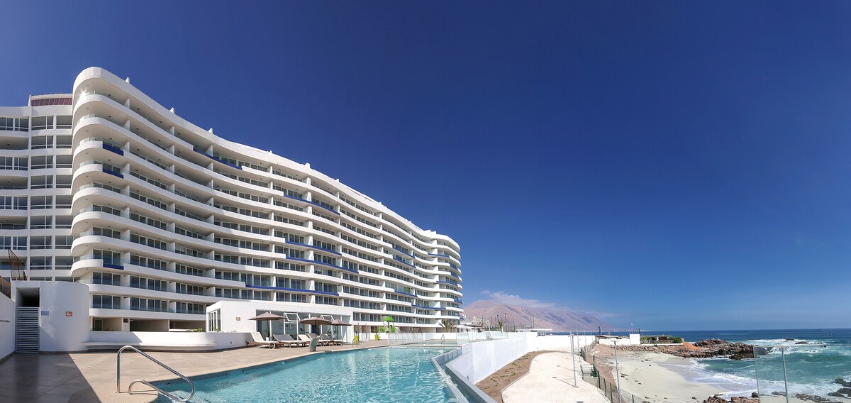 An expansive view showcases a modern building along the coastline, with multiple balconies facing the ocean. A shimmering pool is positioned in the foreground, reflecting the clear blue sky, while sandy beach and ocean waves are visible in the background.
