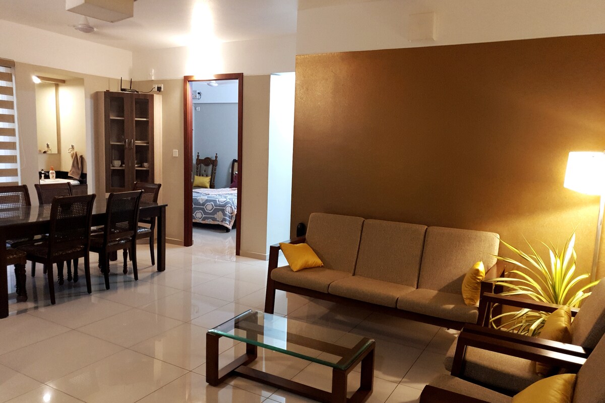 The living area is furnished with a modern sofa accompanied by vibrant cushions. A glass coffee table sits atop shiny white tiles. In the background, a dining table with wooden chairs is visible, along with a glimpse of a well-lit room through an open doorway.