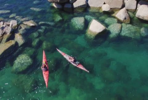 Two kayakers are seen navigating through clear turquoise waters, surrounded by smooth, natural rock formations. The water reveals a rocky floor, enhancing the vibrant color palette of the scene.