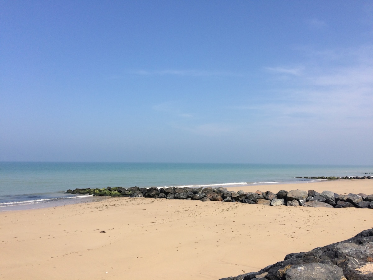 A sandy beach extends to meet the calm blue sea under a clear sky. Smooth stones line the shoreline, providing a natural barrier. Gentle waves lap at the water's edge, and patches of green algae are visible on the rocks.