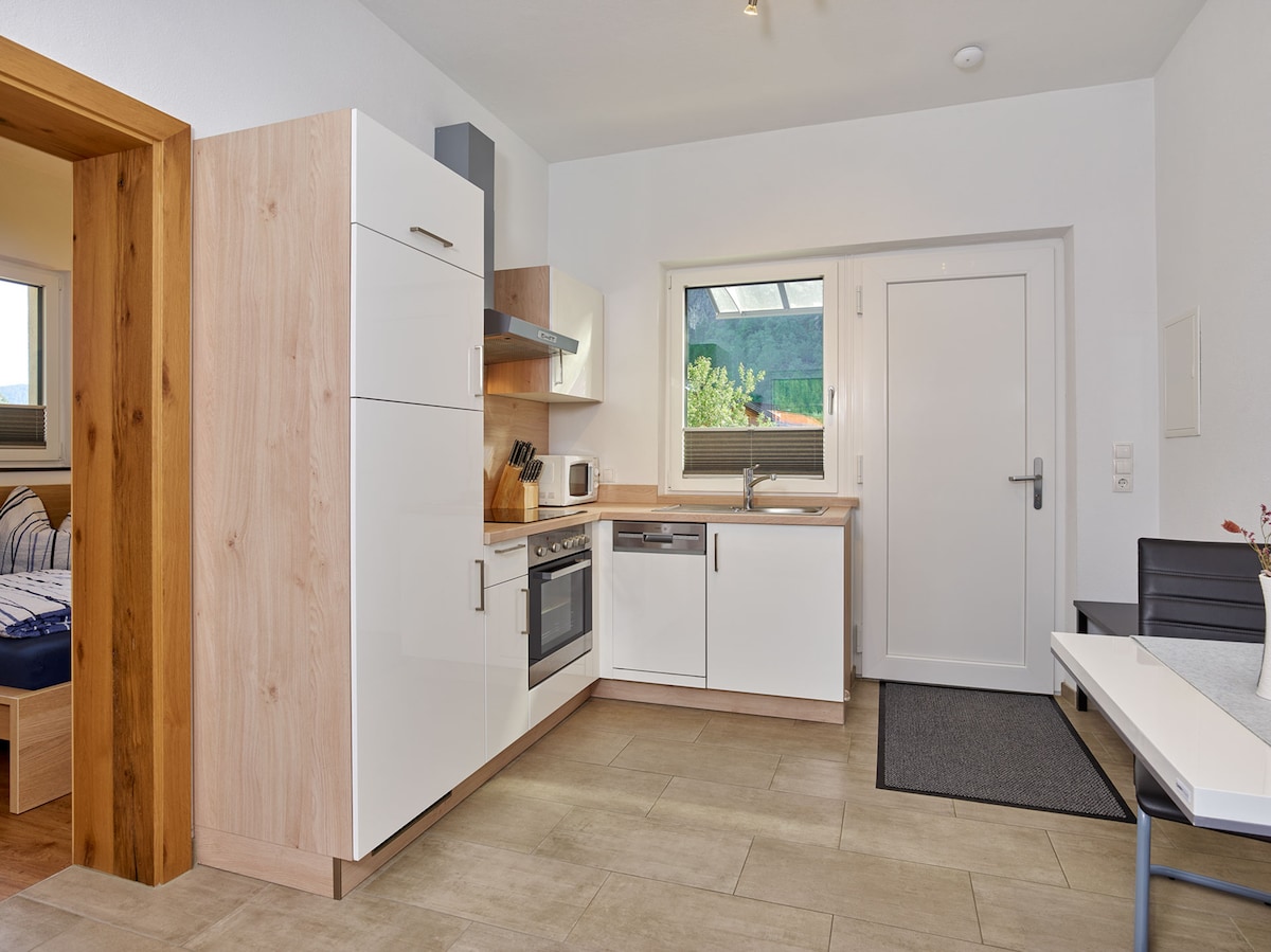 A bright kitchen area is visible, featuring modern white cabinetry and stainless steel appliances. A sink, stovetop, and microwave are included. Natural light enters through a window, illuminating the tile flooring. A dining table is present in the adjacent space, enhancing functionality.