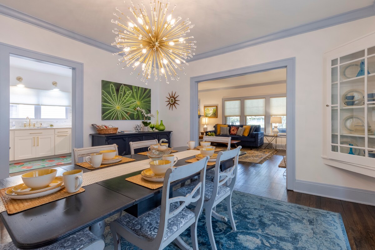 A dining area is presented with a dark wooden table set for six, featuring plates and cups in an organized arrangement. A modern chandelier hangs overhead, illuminating the space. Decor accents include a vibrant painting and a cozy living room visible through the adjacent archway.