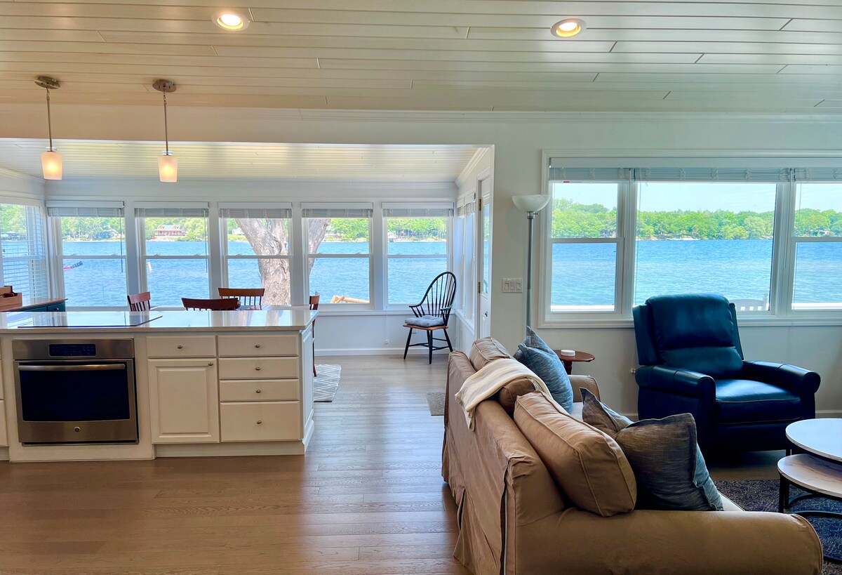 A bright and airy great room is visible, featuring large windows that offer views of the water. The space includes a comfortable sofa and a recliner, with subtle highlights of wood in the dining area and nearby seating. Soft light illuminates the interior from multiple sources.