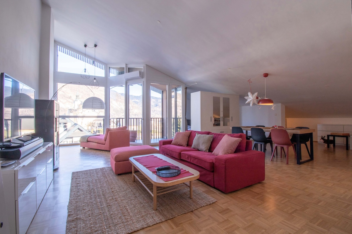 The spacious living area features modern furnishings in neutral and vibrant tones, including a pink sofa and a low coffee table. Large windows provide a panoramic view of the mountains, allowing natural light to fill the space. A dining area with a dark table and colorful chairs is visible in the background.