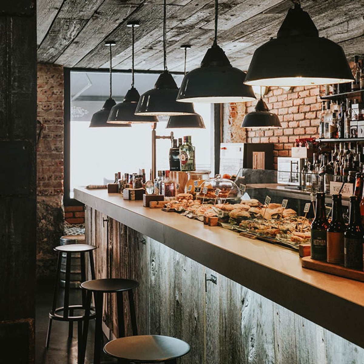 A rustic bar area is highlighted by a long wooden countertop, adorned with an assortment of small plates and beverages. Industrial-style pendant lights hang above, casting warm light over the space. The atmosphere conveys a casual dining experience with a focus on local flavors.