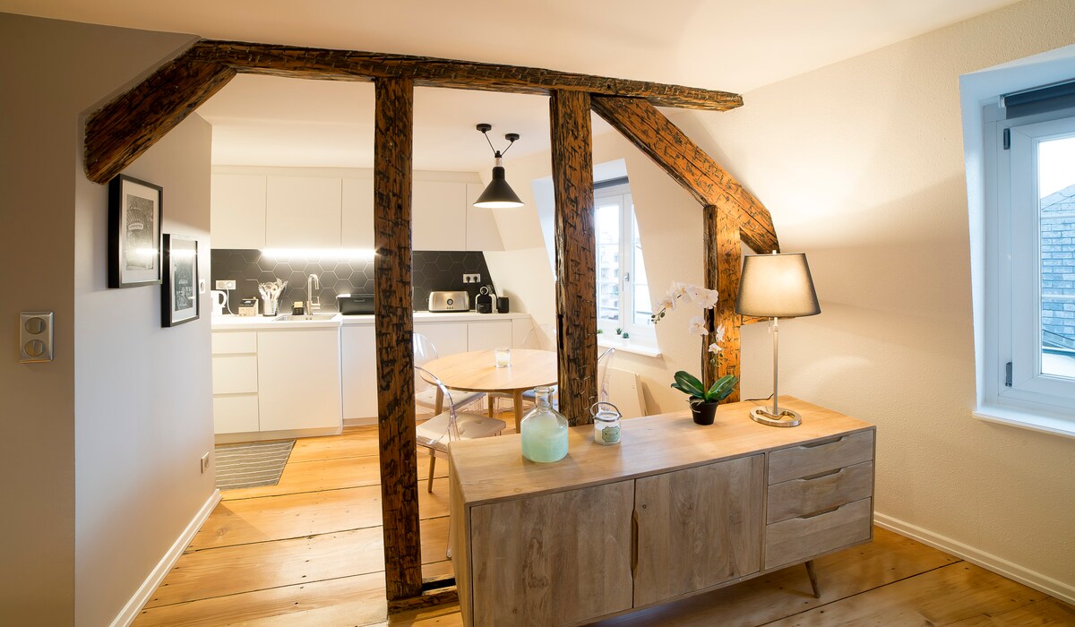 Wooden beams create a distinct architectural detail above a dining table set for two, located adjacent to a modern kitchen. Natural light illuminates the space, highlighting the clean lines of the furniture and the warm tones of the wooden floor.
