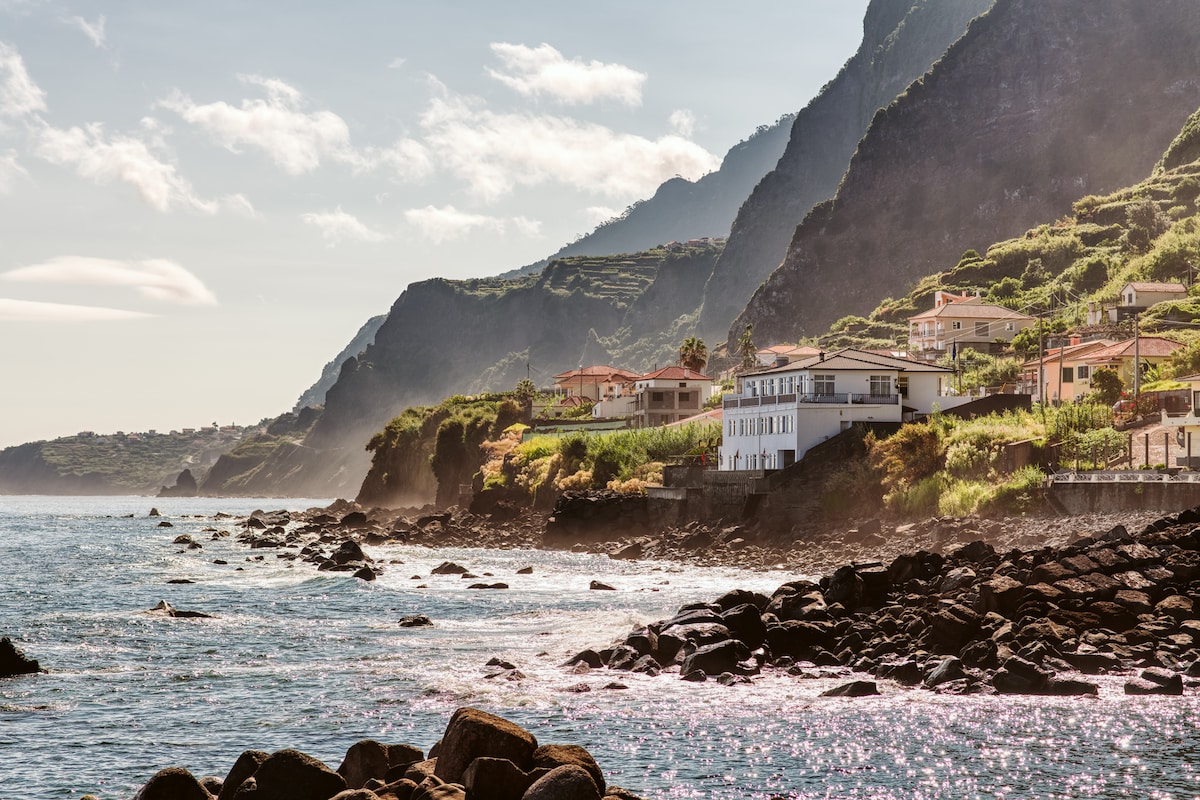 A scenic view of the coastline is captured, featuring rugged rocks along the water's edge. The backdrop includes terraced hills and residential buildings, with lush greenery cascading down the slopes. Soft sunlight illuminates the scene, creating a serene atmosphere over the ocean.
