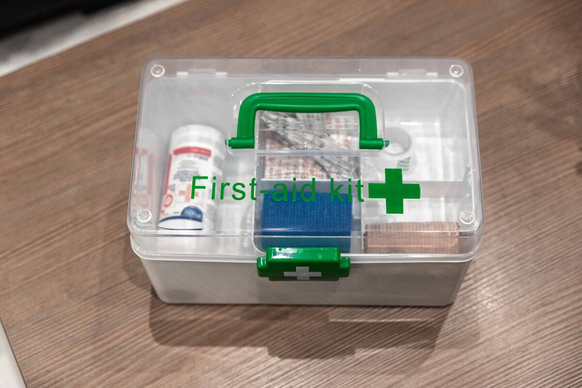 A clear plastic first-aid kit is shown on a wooden surface. The kit features a green handle and a green cross symbol. Inside, various medical supplies, including bandages and medication, are organized neatly, ensuring preparedness for minor emergencies.