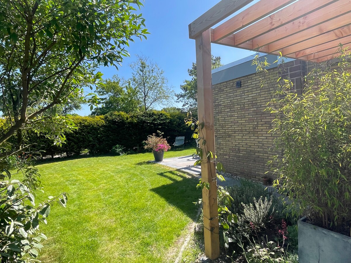 A sunny garden space is presented, featuring well-maintained green grass and a wooden pergola. Flower pots add color to the landscape, while comfortable seating can be seen under the open sky, inviting relaxation. Surrounding greenery provides a sense of privacy and tranquility.