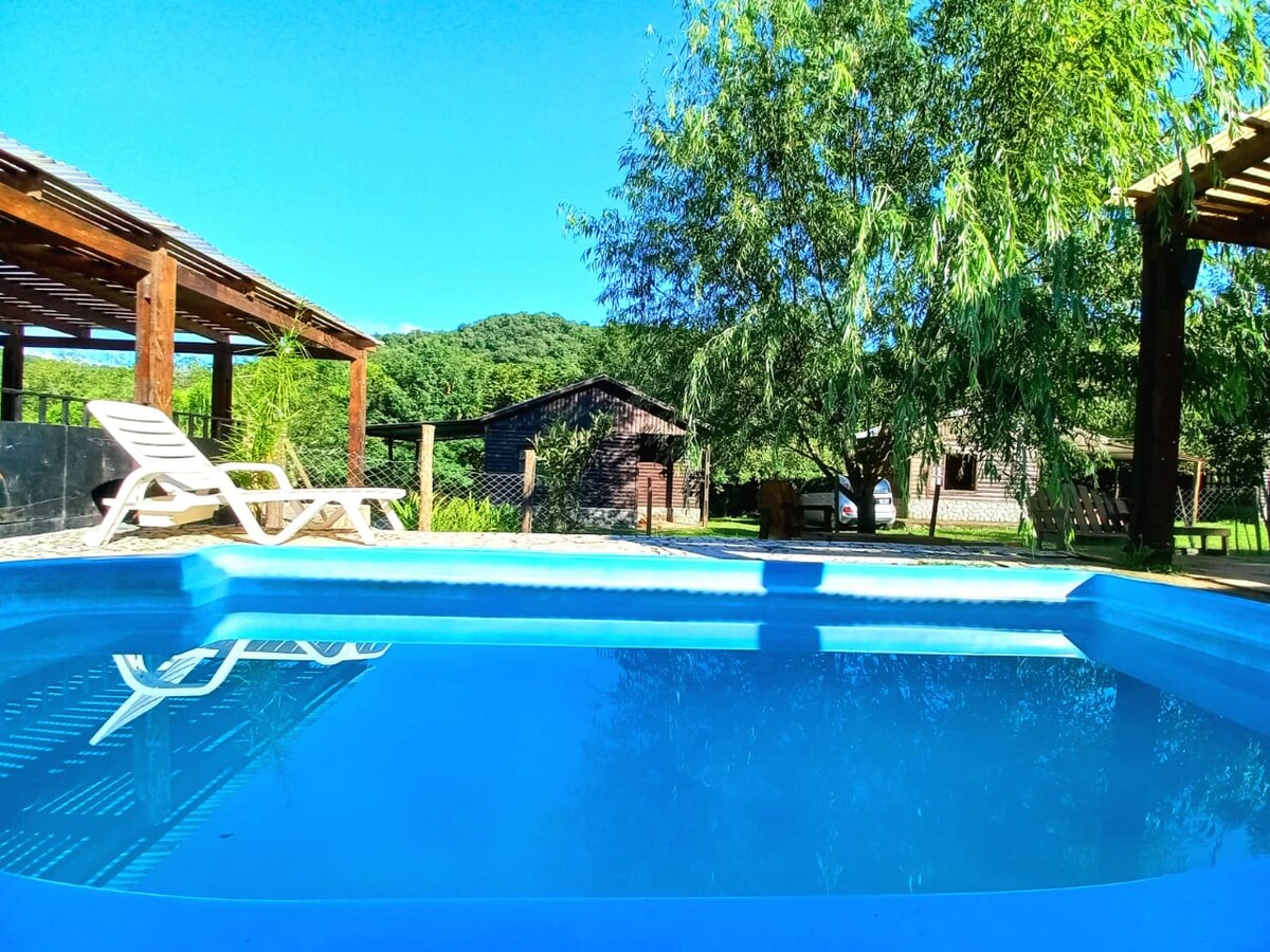 A sparkling pool is visible, surrounded by green foliage and sun loungers. Rustic cabins are seen in the background, along with a wooden pergola. Clear blue skies complement the serene outdoor environment, inviting relaxation and leisure.