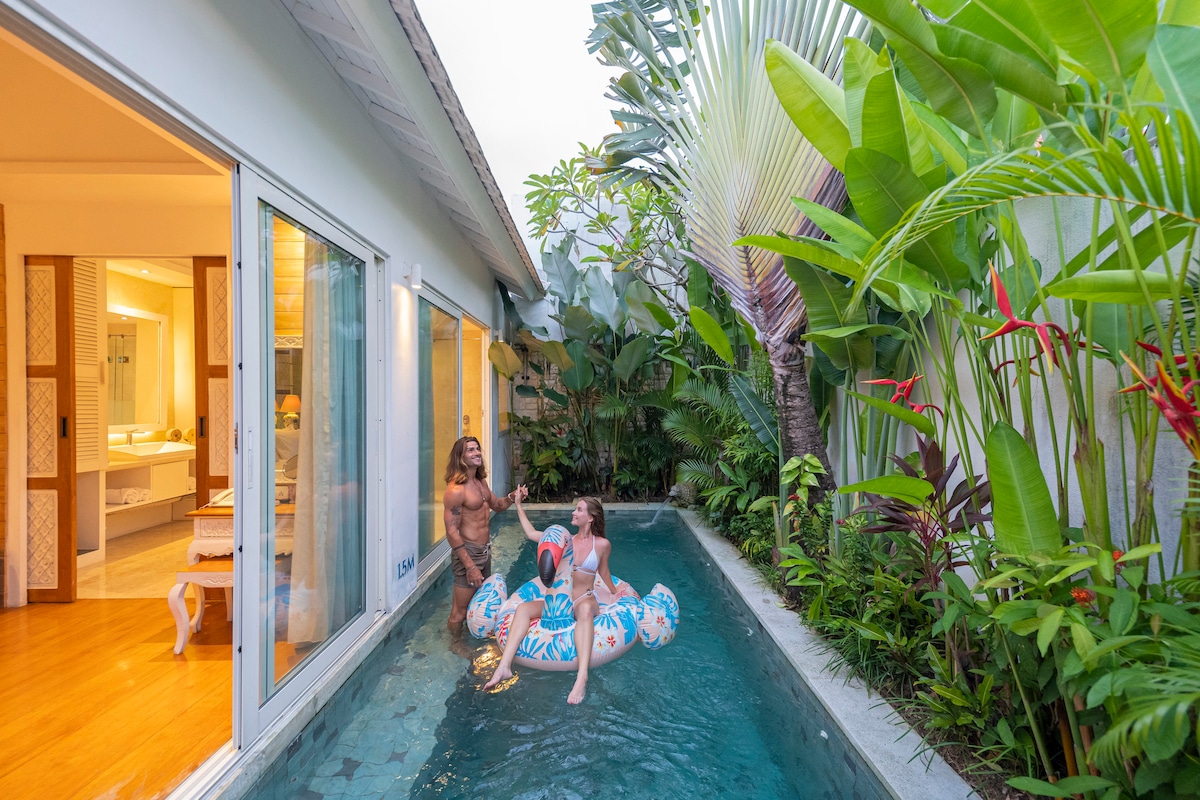 A serene outdoor area features a private swimming pool surrounded by lush tropical plants. The pool is equipped with a float, and sunlight reflects off the water's surface. Glass doors open to a well-lit indoor space, creating a seamless transition between indoor and outdoor settings.