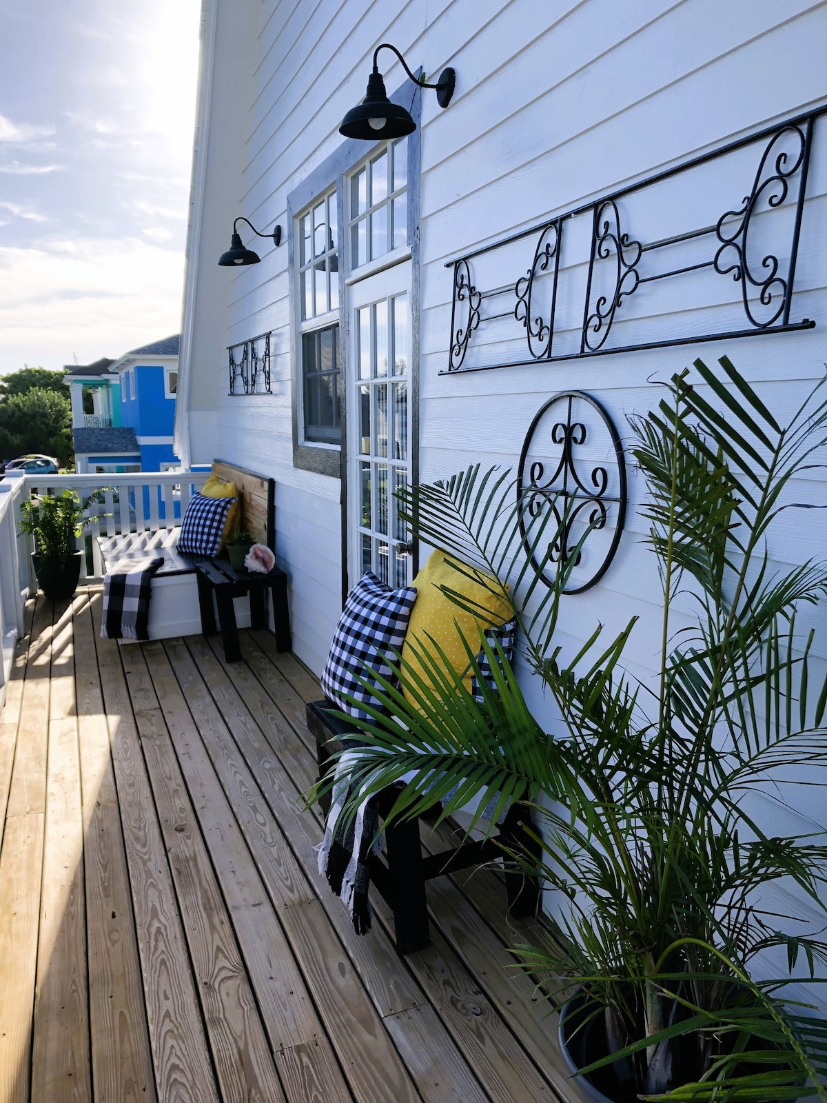 An outdoor reading space features two black chairs adorned with checkered pillows, complemented by vibrant yellow cushions. A small plant adds a touch of greenery, while subtle lighting fixtures are mounted on the white wall beside decorative wall accents.
