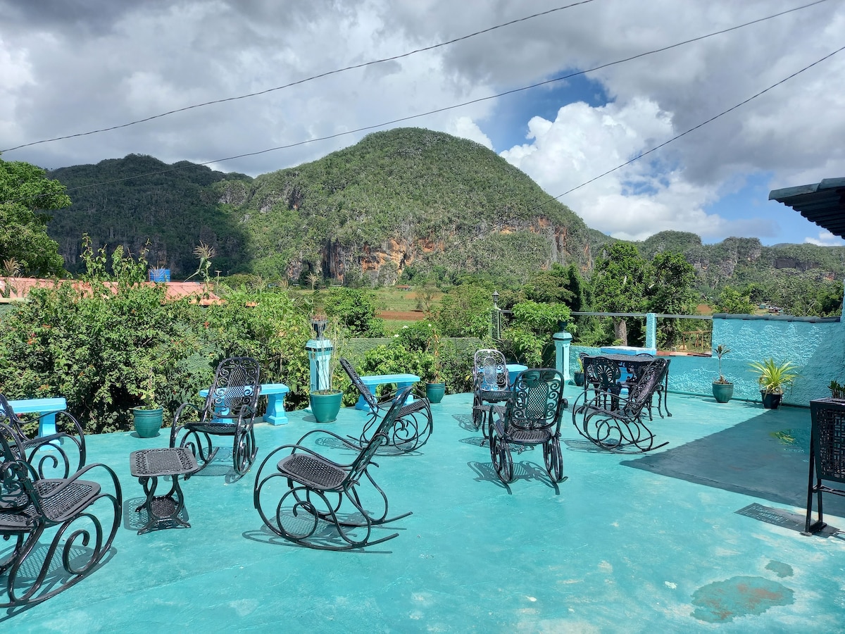 A spacious terrace is observed, featuring several metal tables and chairs arranged for outdoor seating. Lush greenery surrounds the area, and the mountains in the background create a serene setting. The vibrant turquoise floor enhances the inviting atmosphere of the space.