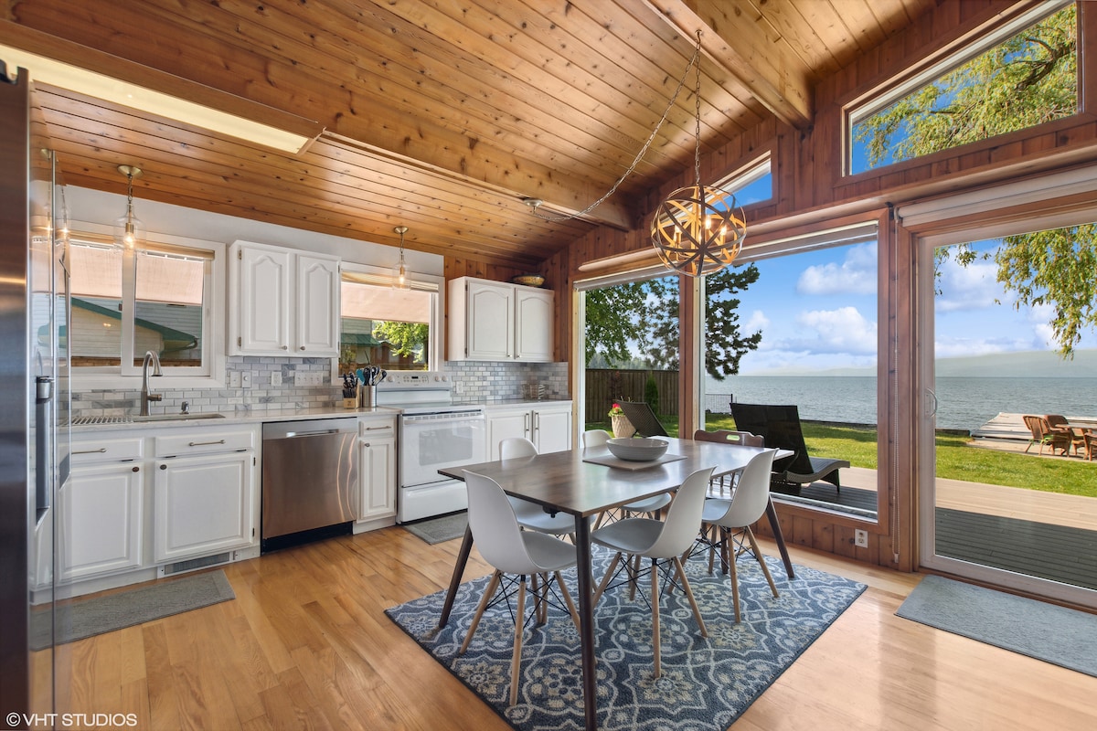The open kitchen and dining area features white cabinetry and modern appliances, with a patterned rug under a dining table surrounded by stylish chairs. Large windows frame a view of the lake, inviting natural light into the space.