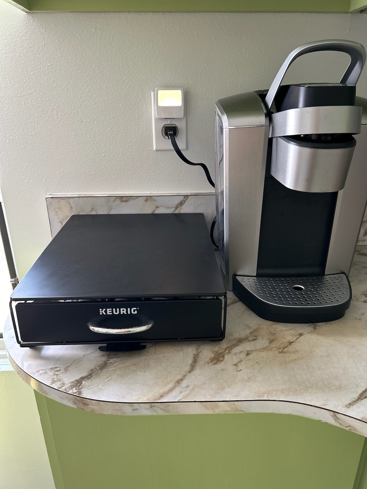 A Keurig coffee maker is positioned on a marble countertop alongside a sleek storage drawer. The coffee maker features a polished design and is plugged into a wall socket, ready for use. Various coffee pods are stored within the drawer, ensuring a selection for morning brews.