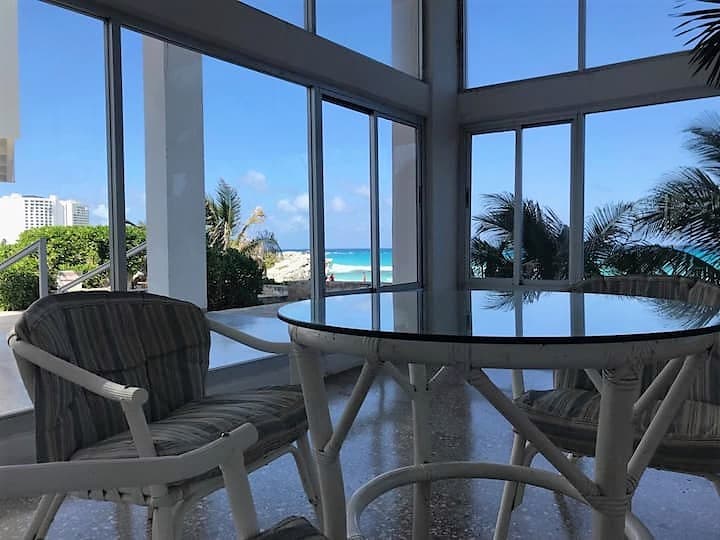 A spacious seating area features a glass table surrounded by striped chairs, with large windows offering a clear view of the turquoise sea and palm trees outside, creating a bright and open atmosphere.