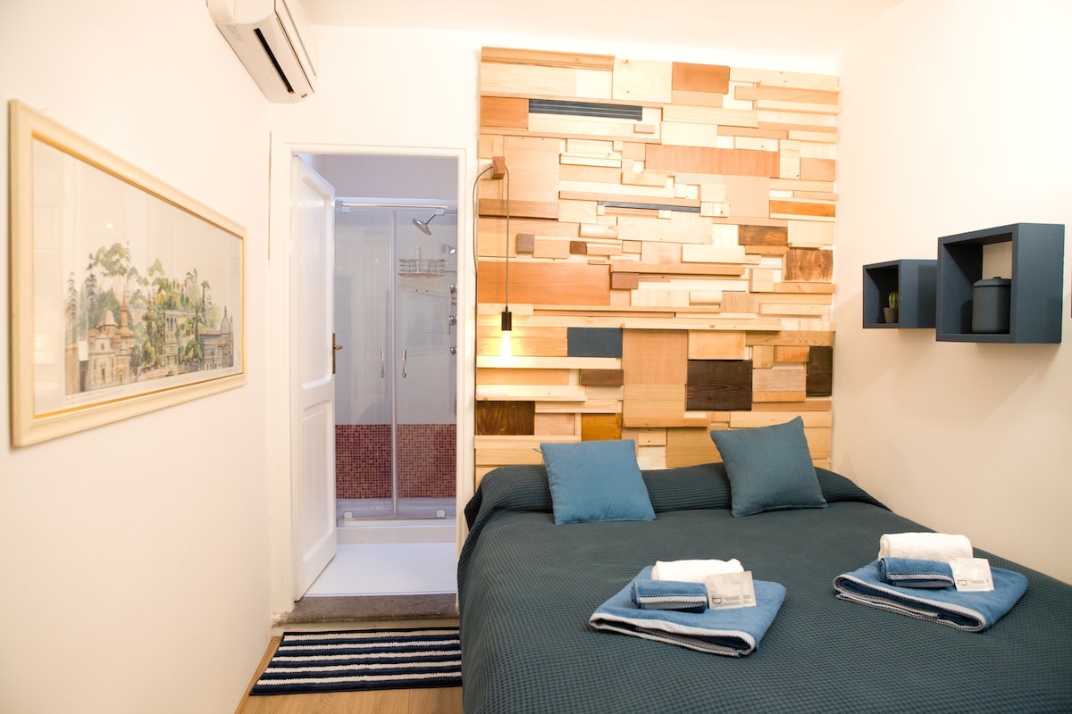 A cozy sleeping area features a spacious bed with blue accent pillows. A textured wooden wall adds warmth to the room. A door leads to a glass-enclosed shower in the bathroom, visible in the background. Soft towels are neatly arranged on the bed.