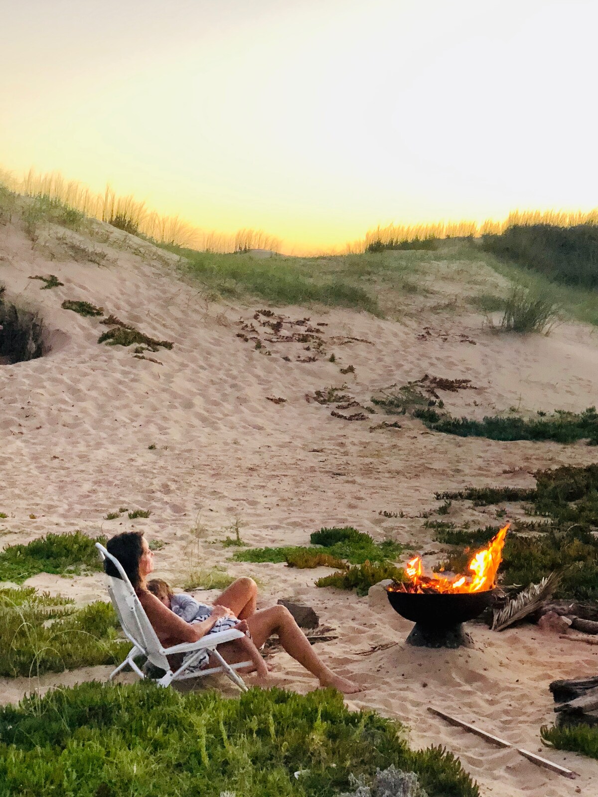 A serene beach setting captures a fire pit surrounded by sandy terrain. A person is seated on a white folding chair, enjoying the warmth of a crackling fire as the sun sets in the background, illuminating the natural landscape.