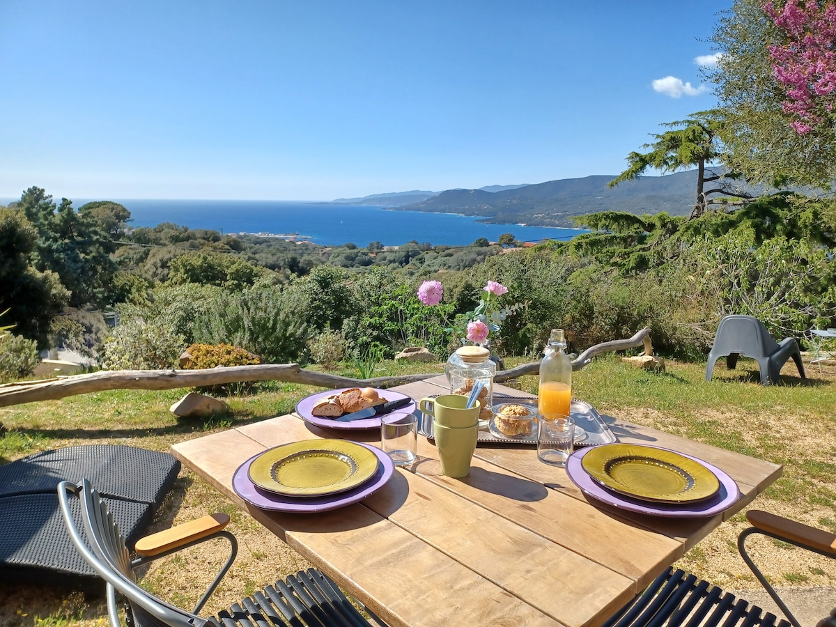 An outdoor dining setting is displayed, featuring a wooden table with purple plates and a glass pitcher beside a bowl of snacks. A scenic view of the coastline is visible in the background, framed by greenery and trees.