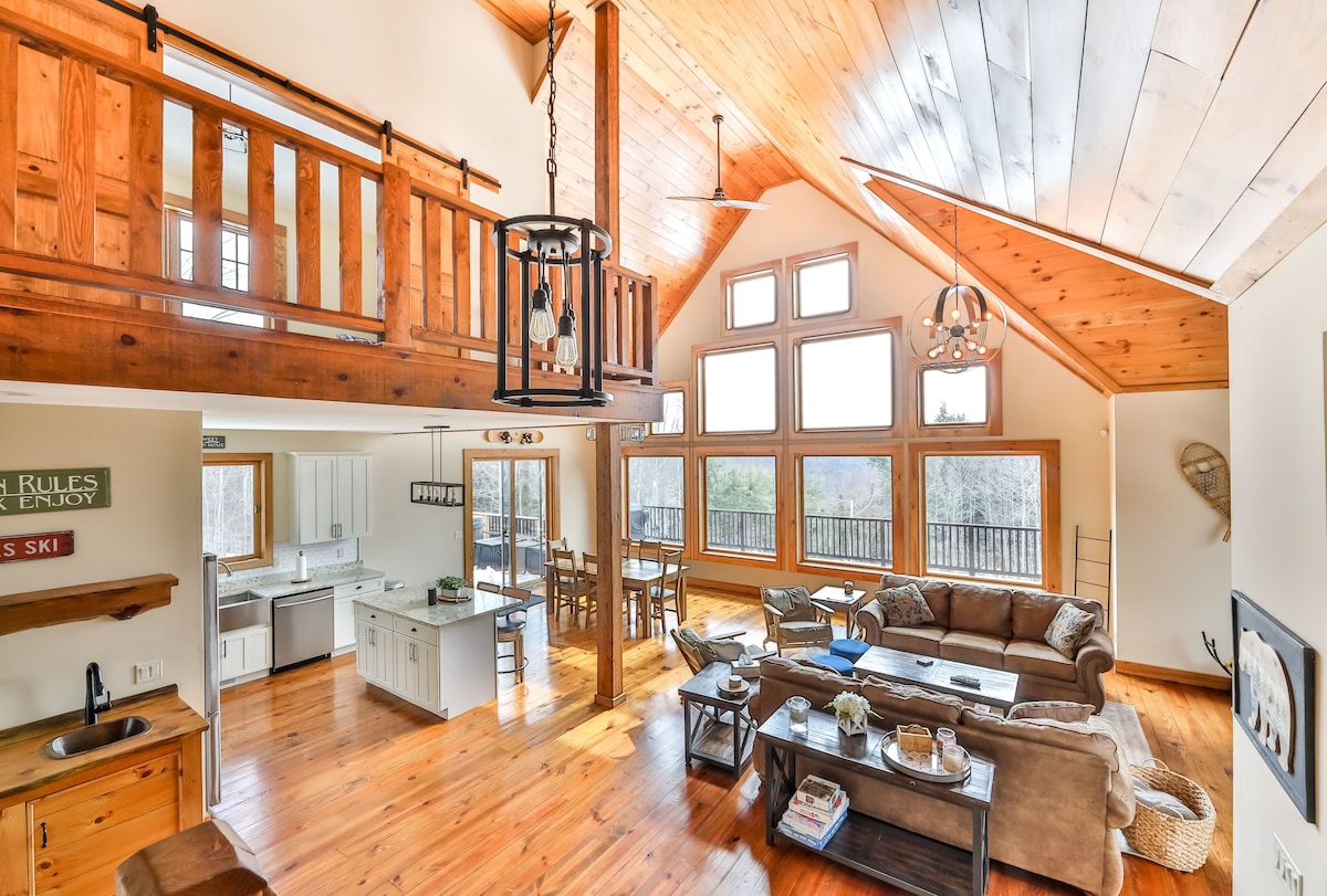 The spacious living area is highlighted by high vaulted ceilings and large windows that allow natural light to fill the space. A cozy sectional sofa is paired with coffee tables, and a dining area is visible in the background, creating an inviting atmosphere for gatherings.