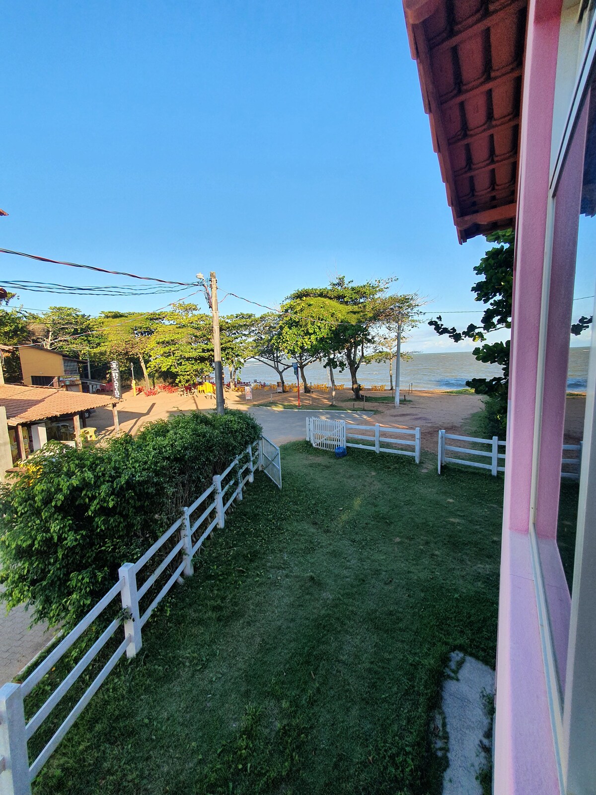 A view from the second floor presents a serene landscape of the beach, framed by lush greenery and trees. A clear path leads towards the ocean, with a white fence marking the property boundary. The sky features a soft blue hue, enhancing the tranquil setting.