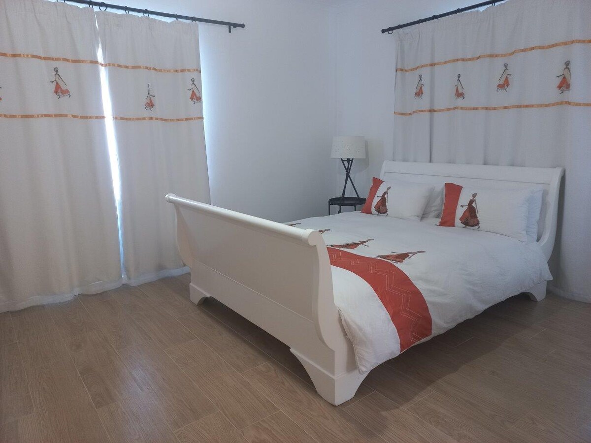 A well-lit bedroom features a queen-sized bed with a minimalistic white frame and decorative orange accents. Soft curtains adorned with a subtle design enhance the room’s ambiance. A bedside lamp and a simple side table contribute to the functional layout, with smooth wooden flooring underfoot.