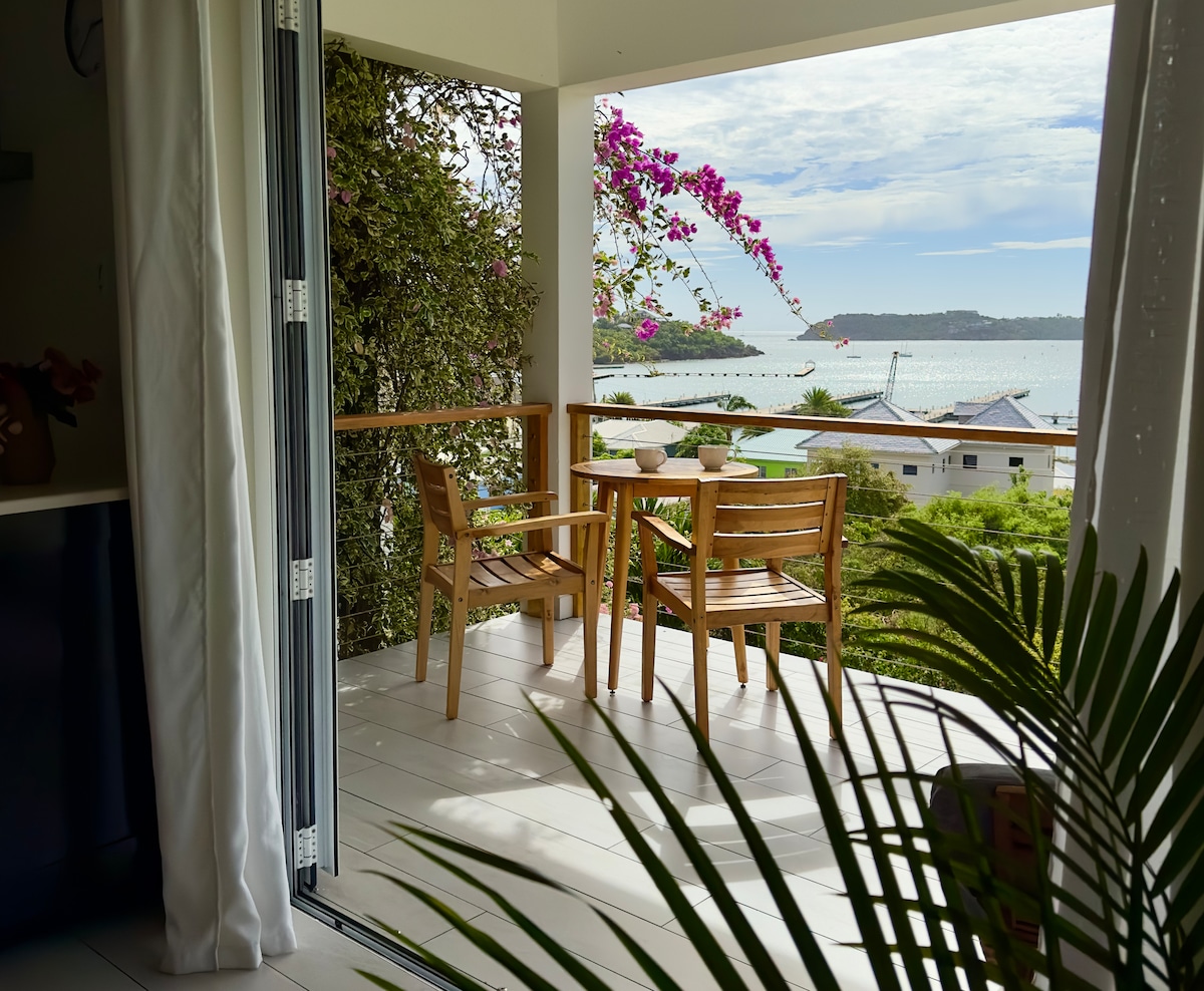 A balcony is visible featuring two wooden chairs and a small table, surrounded by lush greenery and vibrant pink flowers. A scenic view of the bay and distant yachts is framed by the open doors, inviting natural light indoors.
