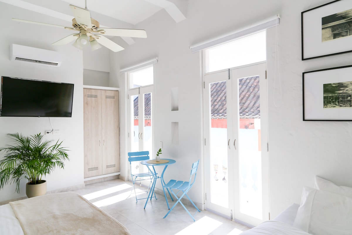 The studio apartment features a bright interior with white walls and natural light streaming through large windows. A cozy seating area consists of a small blue table and two chairs. A television mounted on the wall and a palm plant add to the inviting atmosphere.