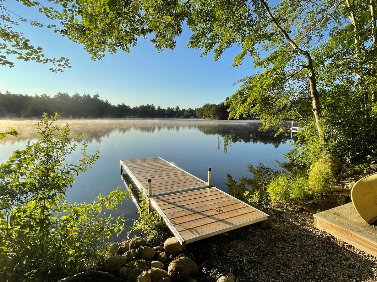 A wooden dock extends over calm waters, surrounded by lush greenery and trees. The early morning mist hovers just above the water's surface, creating a serene atmosphere. The shoreline features smooth stones, inviting guests to enjoy the tranquil lakeside view.