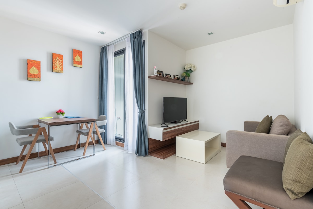 A modern living space features a soft brown couch and a light wooden entertainment unit with a TV. A dining table with four chairs is positioned beside a large window, adorned with flowing curtains. Colorful wall art adds a touch of vibrancy to the neutral walls.
