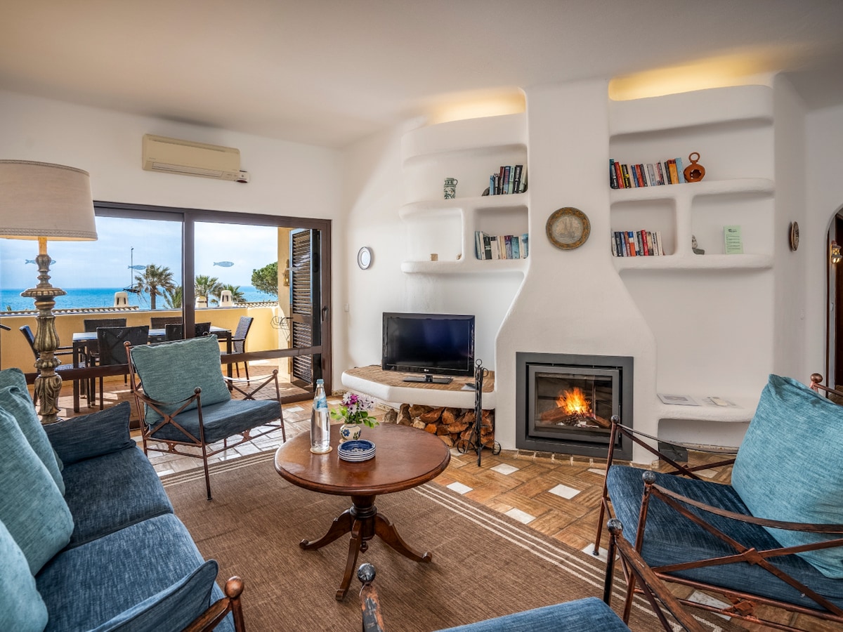 A cozy living room is furnished with blue seating and a round wooden table. A fireplace is positioned against a white textured wall, while built-in shelves display books and decorative items. Large windows offer views of the outdoor terrace, enhancing the room's relaxed ambiance.