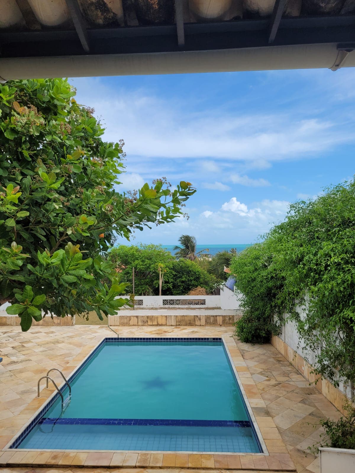 A serene outdoor area features a clean swimming pool with a gentle blue hue, surrounded by natural stone tiles. Lush greenery frames the space, and a glimpse of the ocean is visible in the background under a clear sky.
