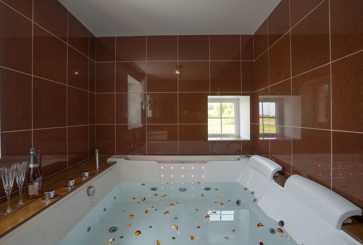 A modern balneo space is featured, with a spacious hot tub filled with water and scattered rose petals. The walls are accented with rich brown tiles, and a window allows natural light to enter, providing views of the surrounding nature.