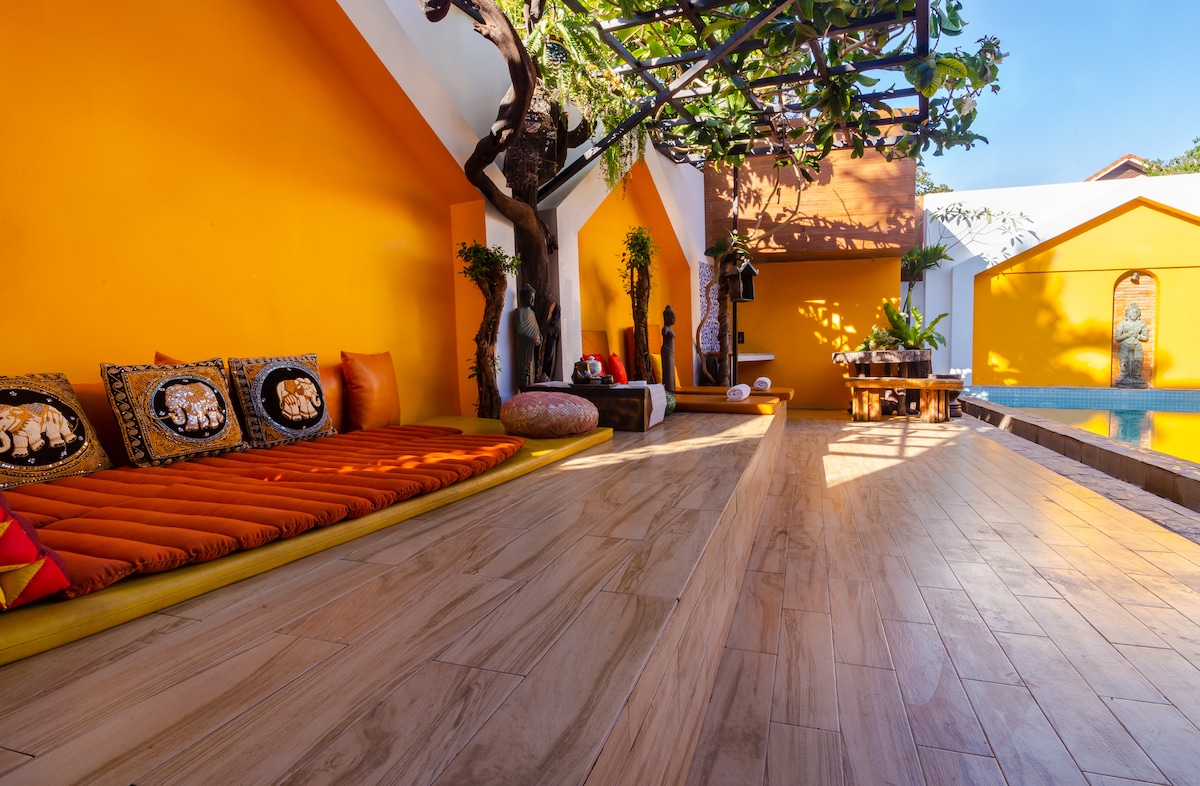 An outdoor sitting area features a wooden deck illuminated by natural light, with an orange canopy overhead. Cozy cushions in vibrant colors are arranged along one side, while potted plants and decorative elements add a touch of greenery. A private pool is visible in the background.