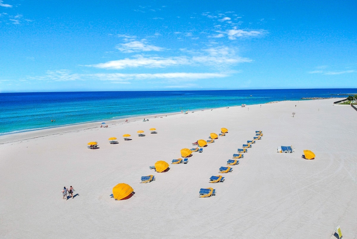 A wide view of a sandy beach showcases a series of yellow beach umbrellas and blue lounge chairs arranged neatly across the shoreline. The vibrant blue water meets the crisp sky, creating a serene coastal atmosphere.