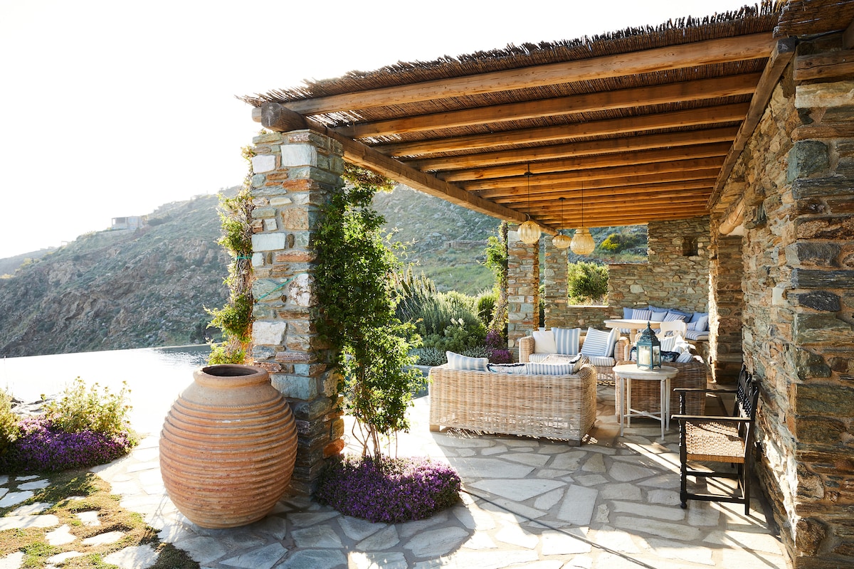 A stone terrace with a wooden ceiling offers a cozy outdoor lounge area. Soft seating is arranged around a low table, framed by colorful flower beds. Natural stone and rustic materials blend harmoniously with the surrounding landscape, providing an inviting space for relaxation.