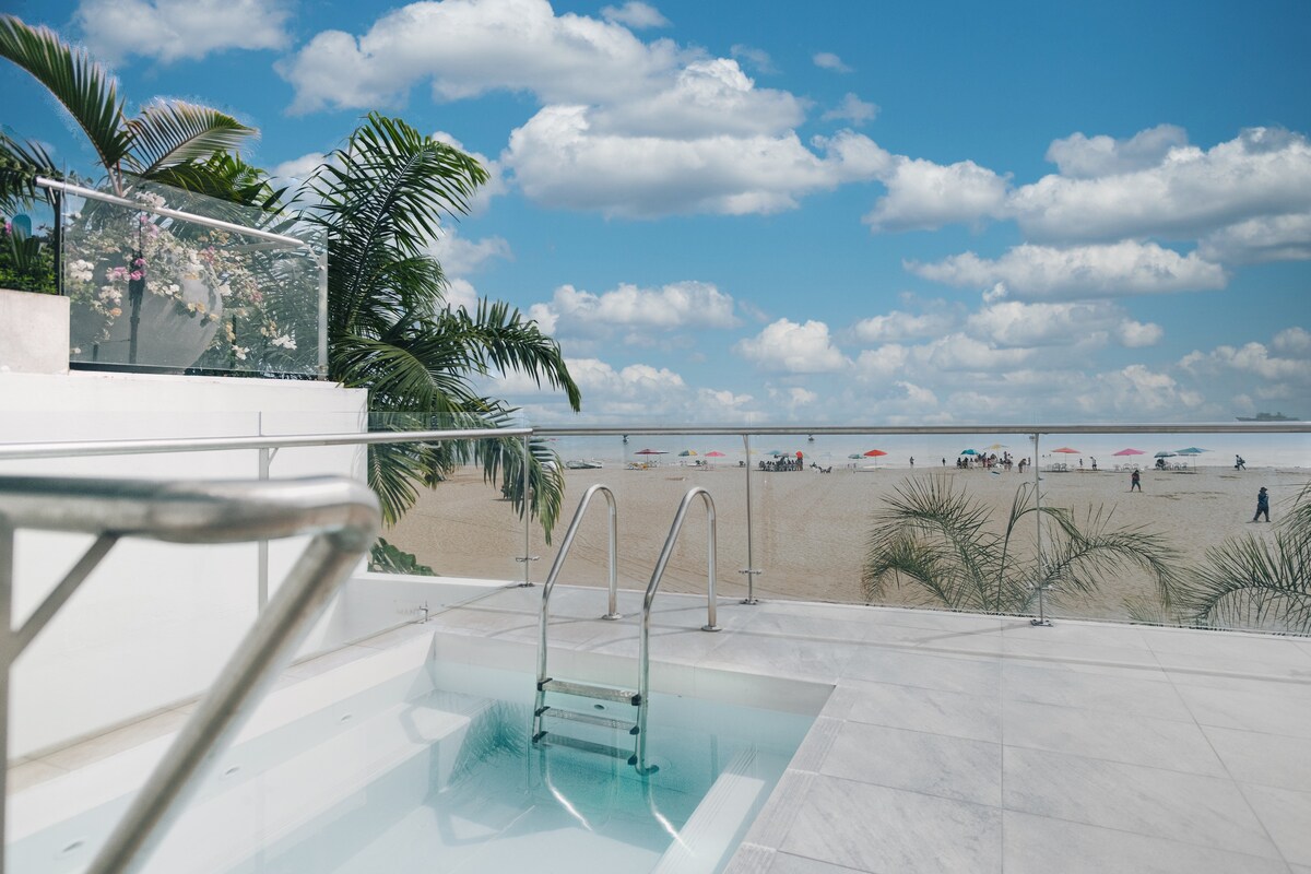 A sleek jacuzzi stands on a modern terrace, surrounded by tropical plants and offering a clear view of the beach. The horizon features gentle clouds and beachgoers enjoying the sandy shore, with colorful umbrellas scattered across the landscape.