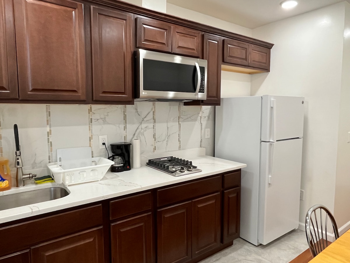 The kitchen features dark wooden cabinetry and a light-colored countertop. A stainless-steel microwave and a large refrigerator are incorporated, alongside a gas stove. A sink with a dish rack is present, and neutral-colored tile backsplashes add a modern touch.