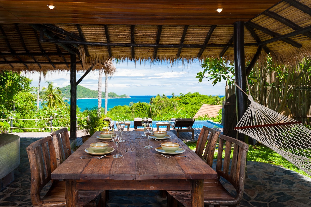 A wooden dining table is set for a meal, featuring tableware and glasses. Behind it, a serene view of the ocean and lush greenery is visible, while a hammock is suspended nearby, inviting relaxation. The area is shaded by a thatched roof, enhancing the tranquil atmosphere.