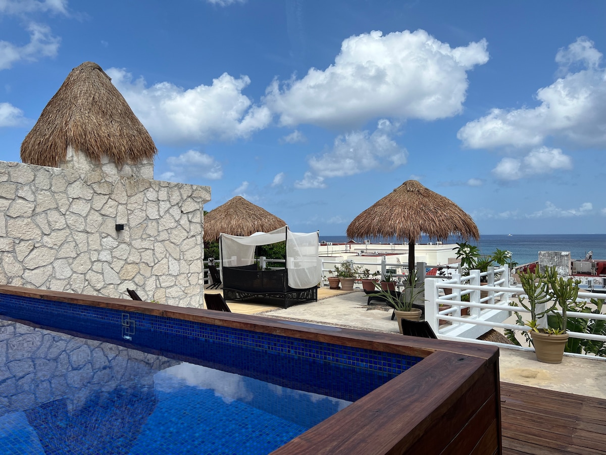 A rooftop area features a rectangular swimming pool with azure tiles, surrounded by wooden decking. Thatched umbrellas offer shade over lounge chairs. A stony wall provides privacy, while the horizon displays the blue sky dotted with fluffy clouds and distant views of the waterfront.