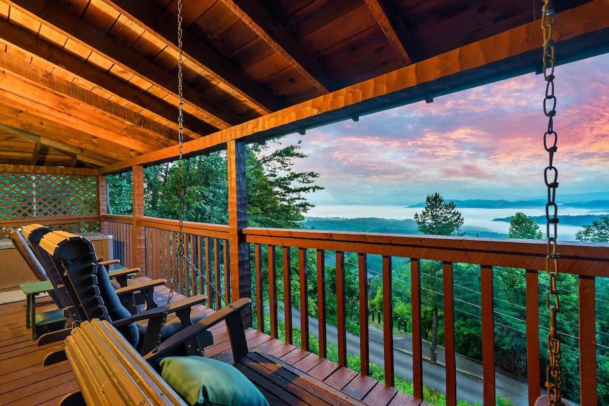 A spacious porch features wooden rocking chairs, offering a comfortable space to enjoy the mountain views. The surrounding railing is crafted from natural wood, complementing the serene environment. The colorful sky at sunset casts a gentle glow over the landscape.